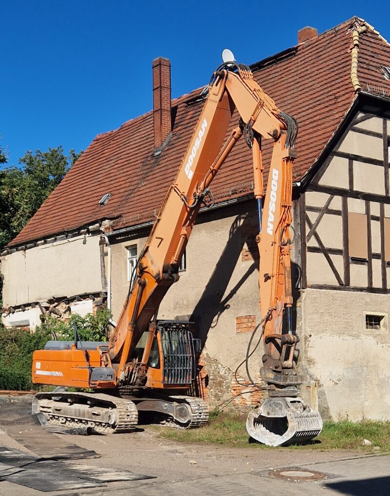 Ein Abrissbagger steht neben einem Fachwerkhaus.